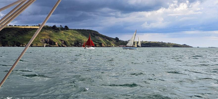 View from a boat showing two sailboats, one with red sails, on choppy grey-green water. Hilly green coastline under cloudy sky in background.